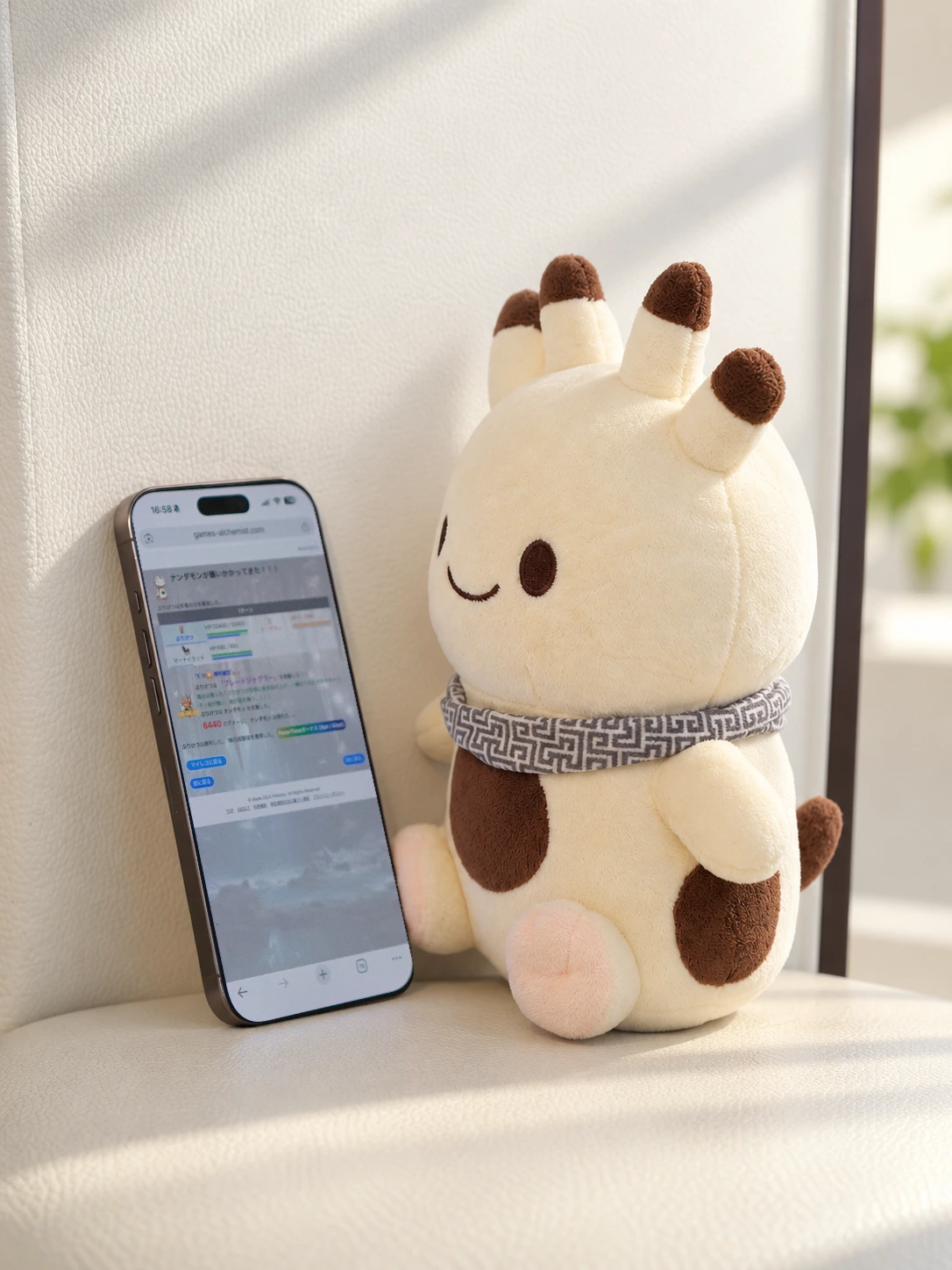 白い椅子の上で、スマートフォンの隣に座るナンダモンのぬいぐるみ。スマートフォンには、あるけみすとでナンダモンと戦う画面が映っている。